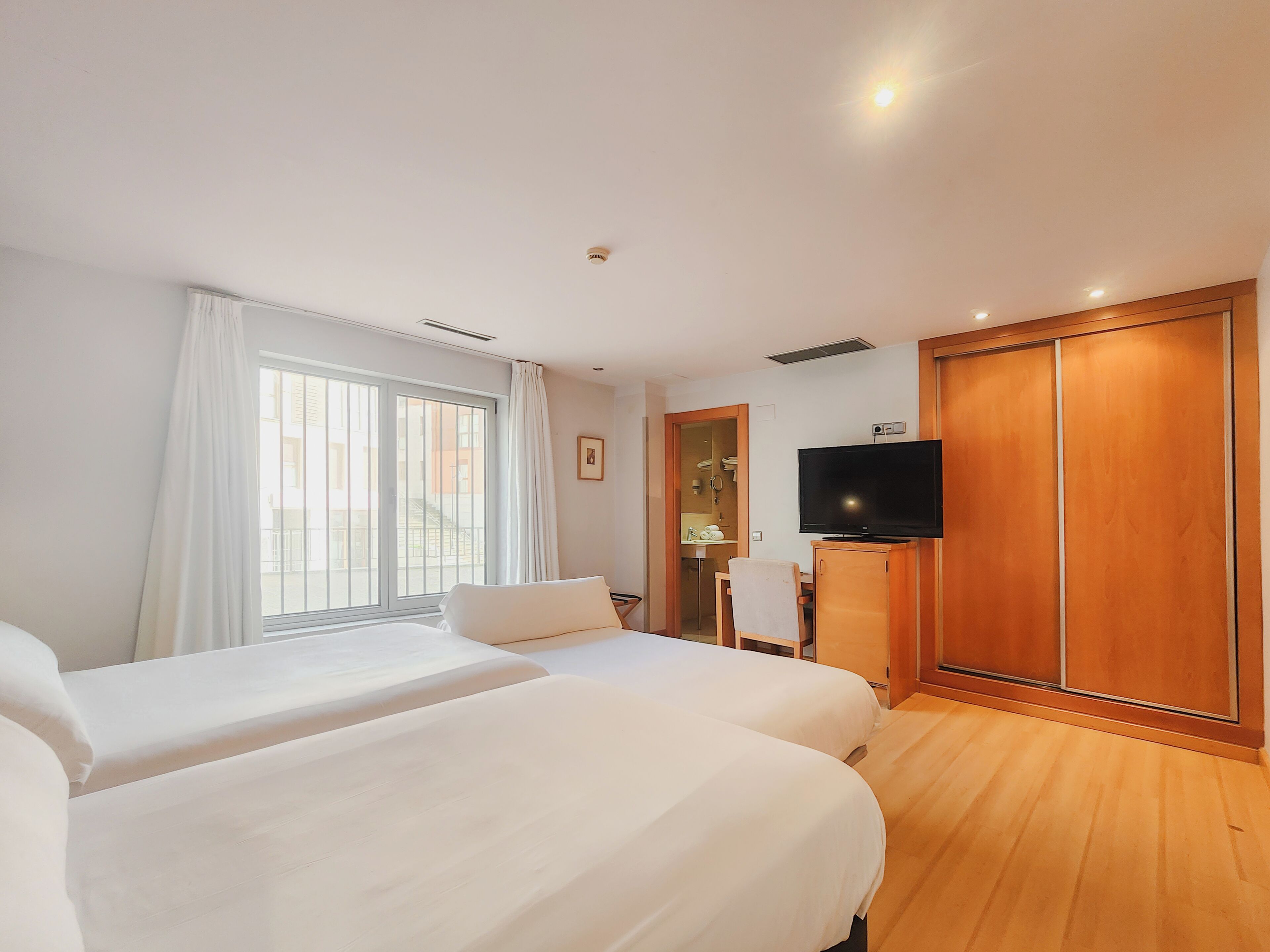 A hotel room with two beds, a TV, a wooden wardrobe, and a bathroom visible through a glass door.