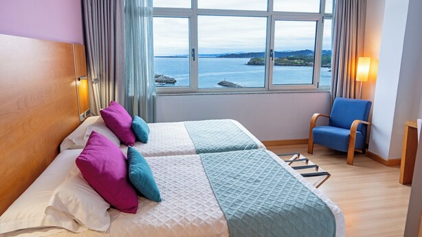 A hotel room with two beds, a view of the sea, a chair, and a lamp.