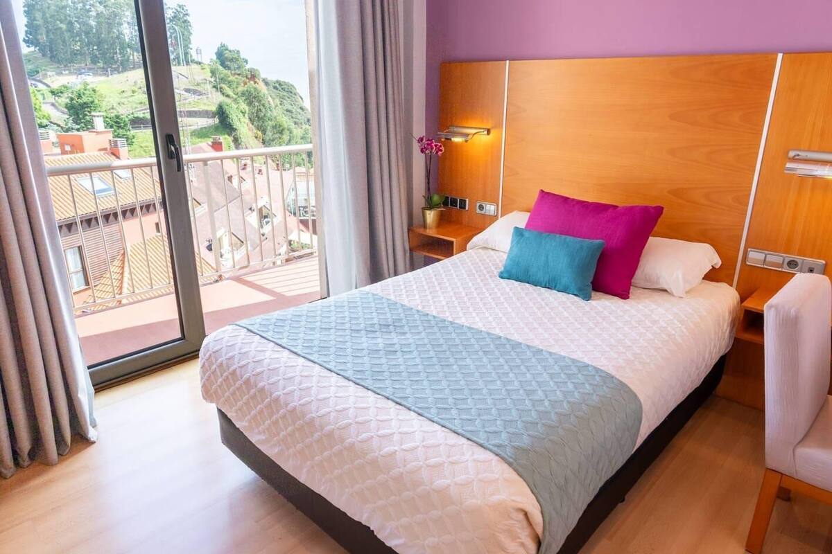 Een hotelkamer met een groot bed, een nachtkastje, een lamp en uitzicht op een balkon met een balustrade.