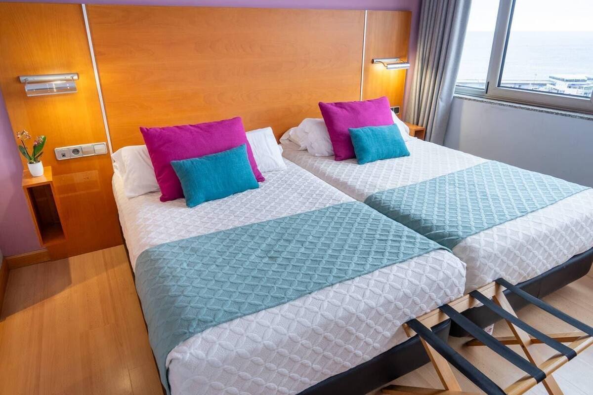 Een hotelkamer met twee bedden, elk voorzien van een gewatteerde spreidek en kussens, een houten hoofdbord, een nachtkastje met een lamp, en uitzicht op zee door een raam.