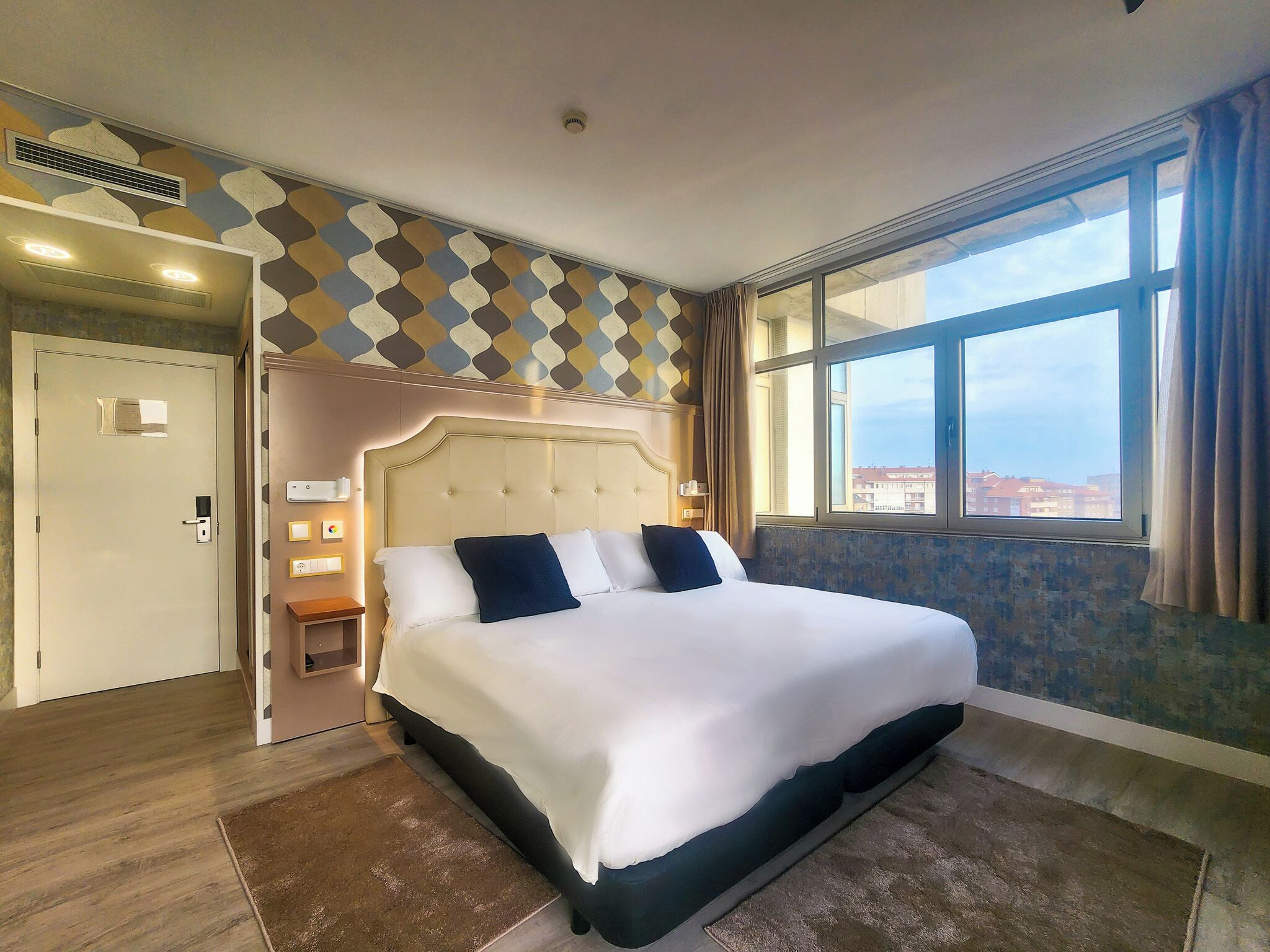 A hotel room with a large bed, a window with a view, a door, and a patterned wall.