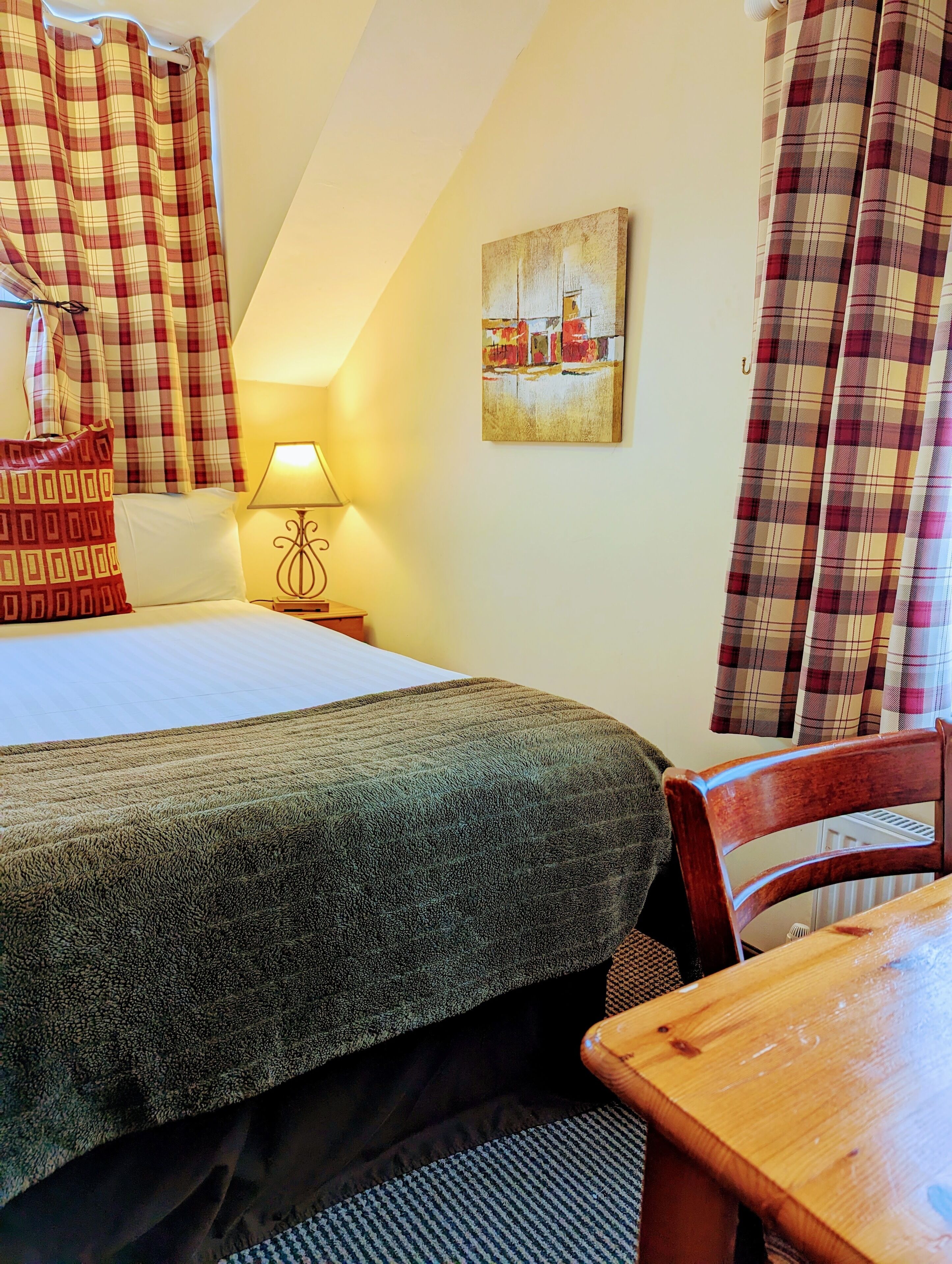 A hotel room with a bed, a table, a chair, a lamp, and curtains.
