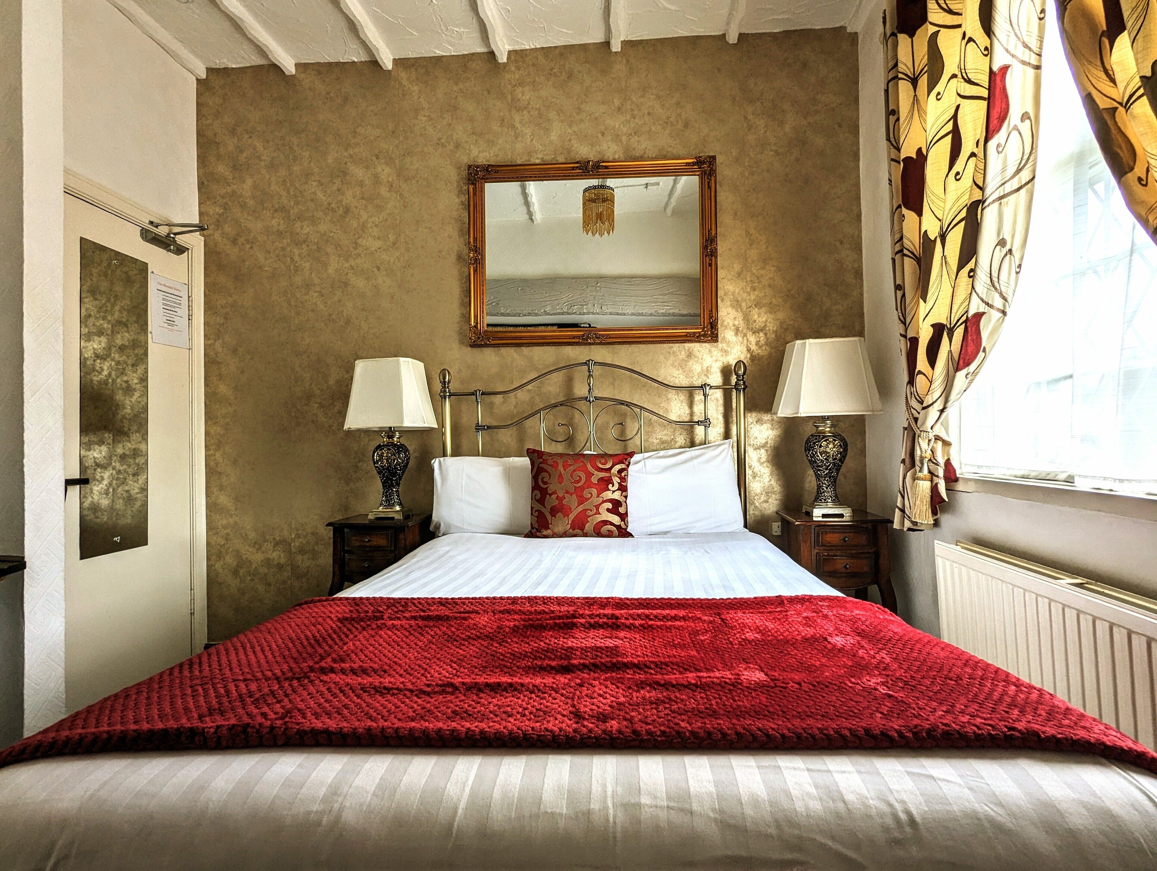A neatly made bed with a red blanket, two bedside tables with lamps, a mirror above the bed, and a window with curtains.