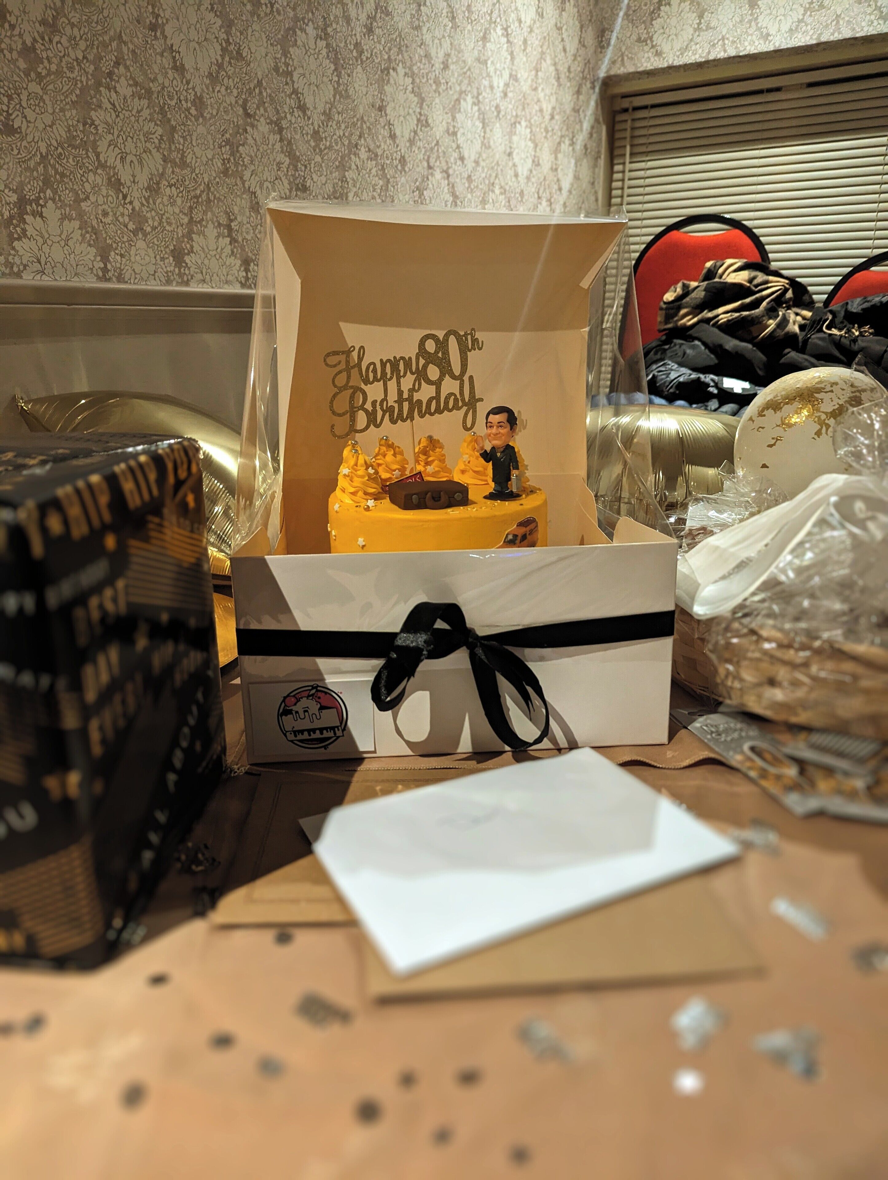 A birthday cake with a 'Happy 80th Birthday' topper in a box, surrounded by various items on a table.