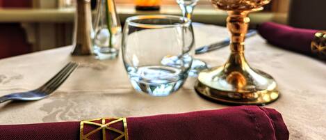 Una mesa con una copa de cristal, un candelabro de cristal, un florero de cristal con una rosa roja y una servilleta doblada con un diseño geométrico dorado.