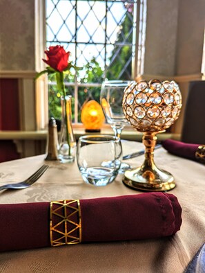 A table set with a glass goblet, a glass candle holder, a glass vase with a red rose, and a folded napkin with a gold geometric design.