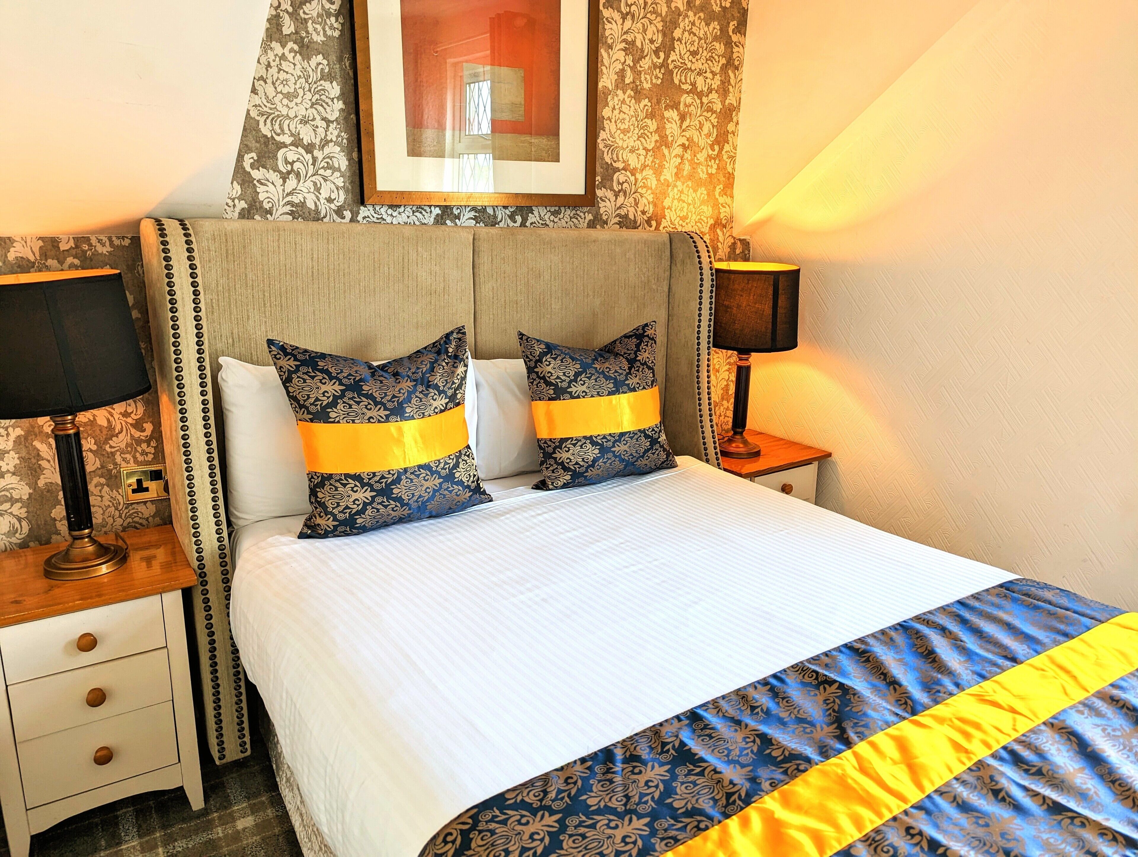 Una cama bien hecha con dos almohadones decorativos, una mesita de noche con lámpara y un cuadro enmarcado en la pared.