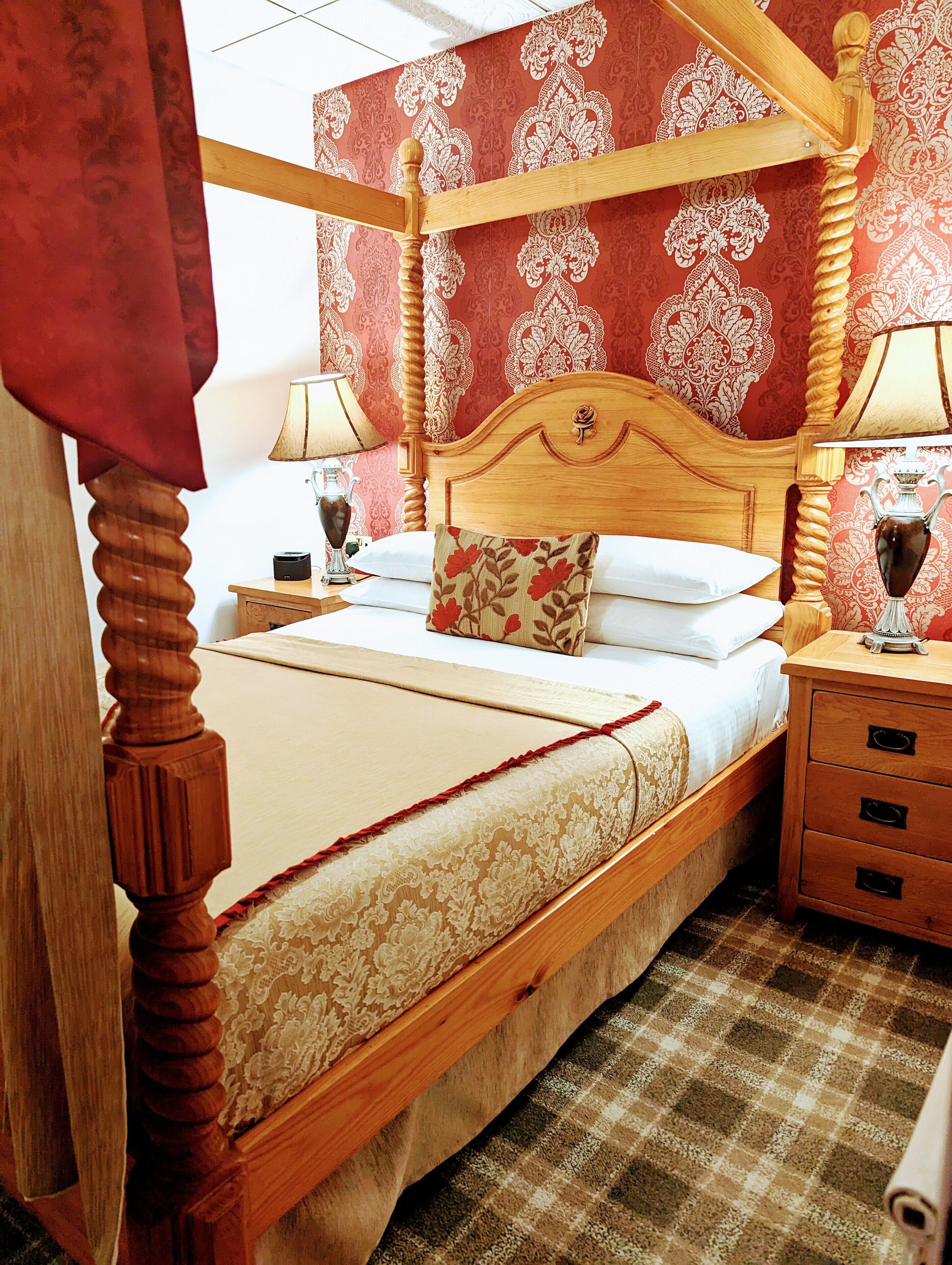 Een hemelbed met houten frame en een hoofdeinde met bloemenprint. Twee nachtkastjes met lampen, een rood gordijn en een geruit vloertje.