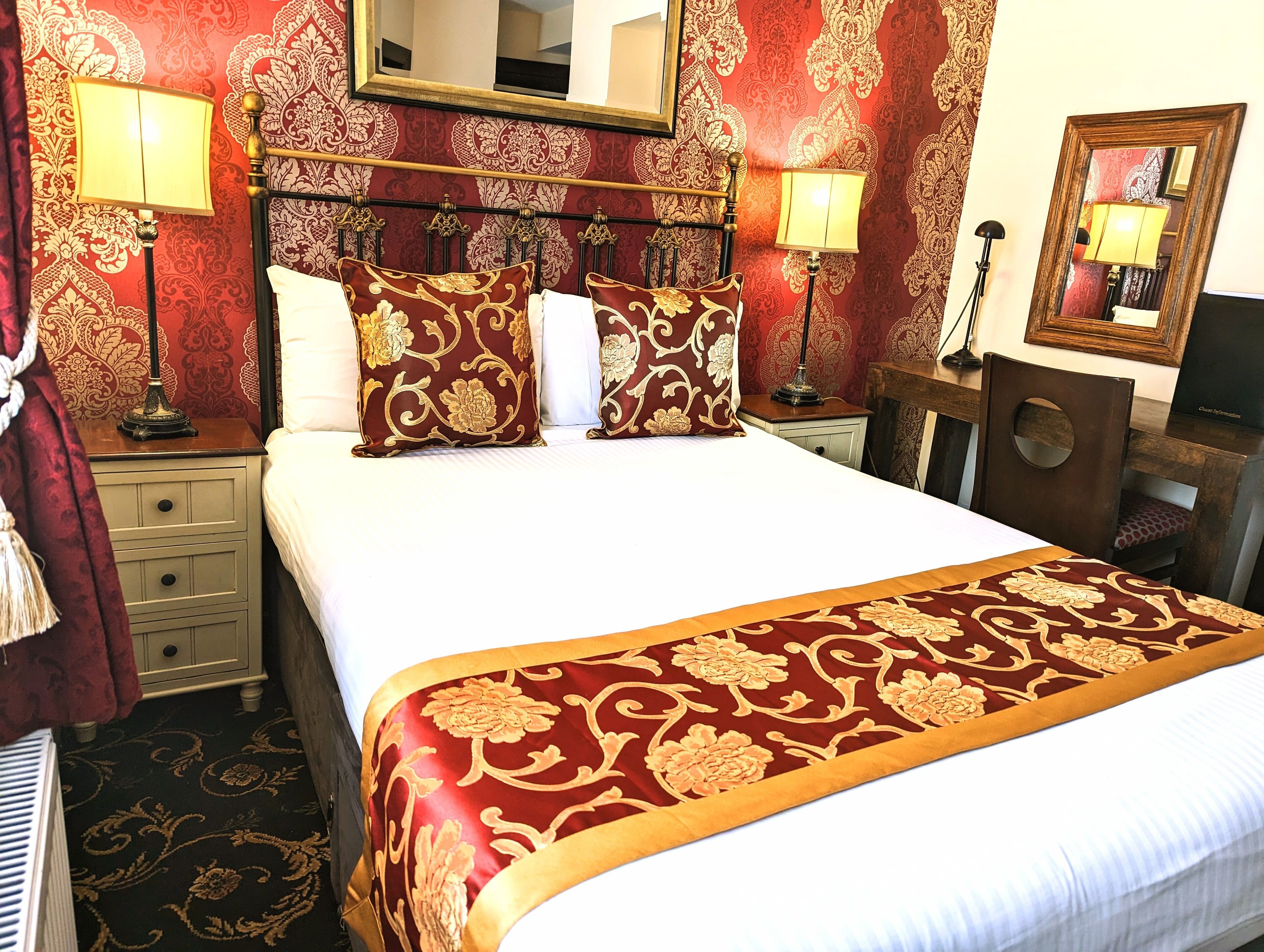 Habitación de hotel con una cama grande, un cabecero decorado, dos mesitas de noche con lámparas, un espejo y una silla de madera.