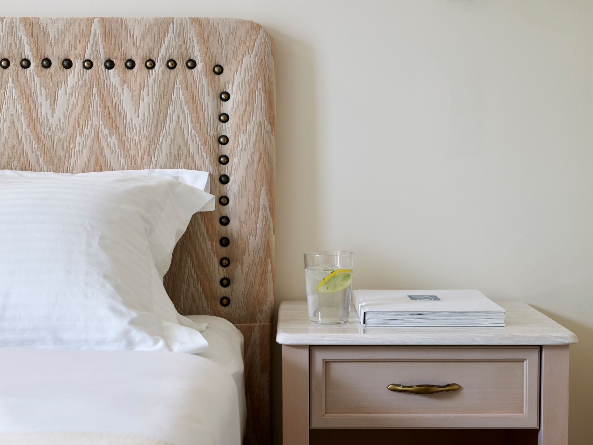 A neatly made bed with white linens, a bedside table with a glass of water and a book, and a drawer with a brass handle.