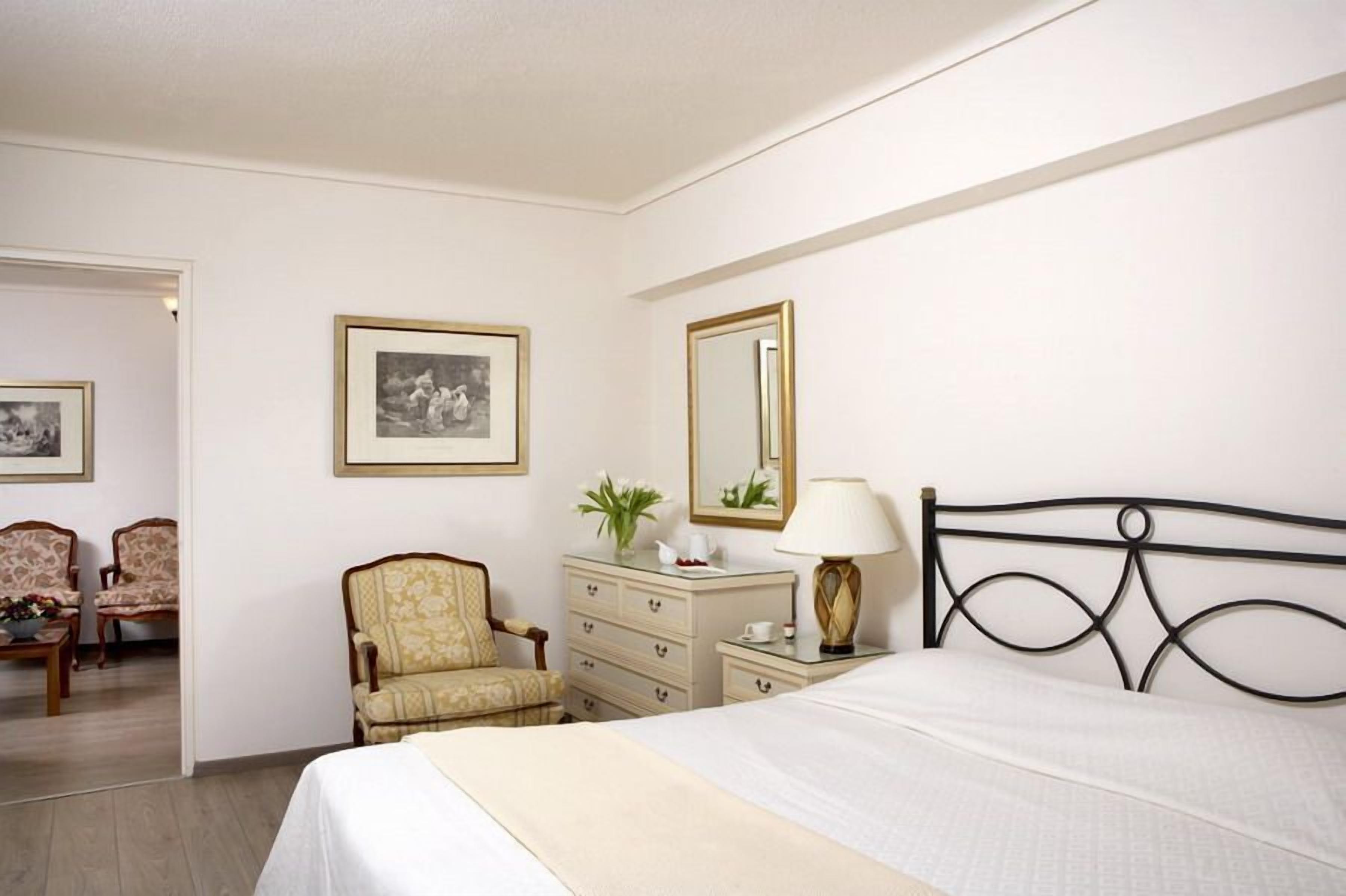 A hotel room with a bed, a dresser, a chair, a mirror, and two framed pictures on the wall.