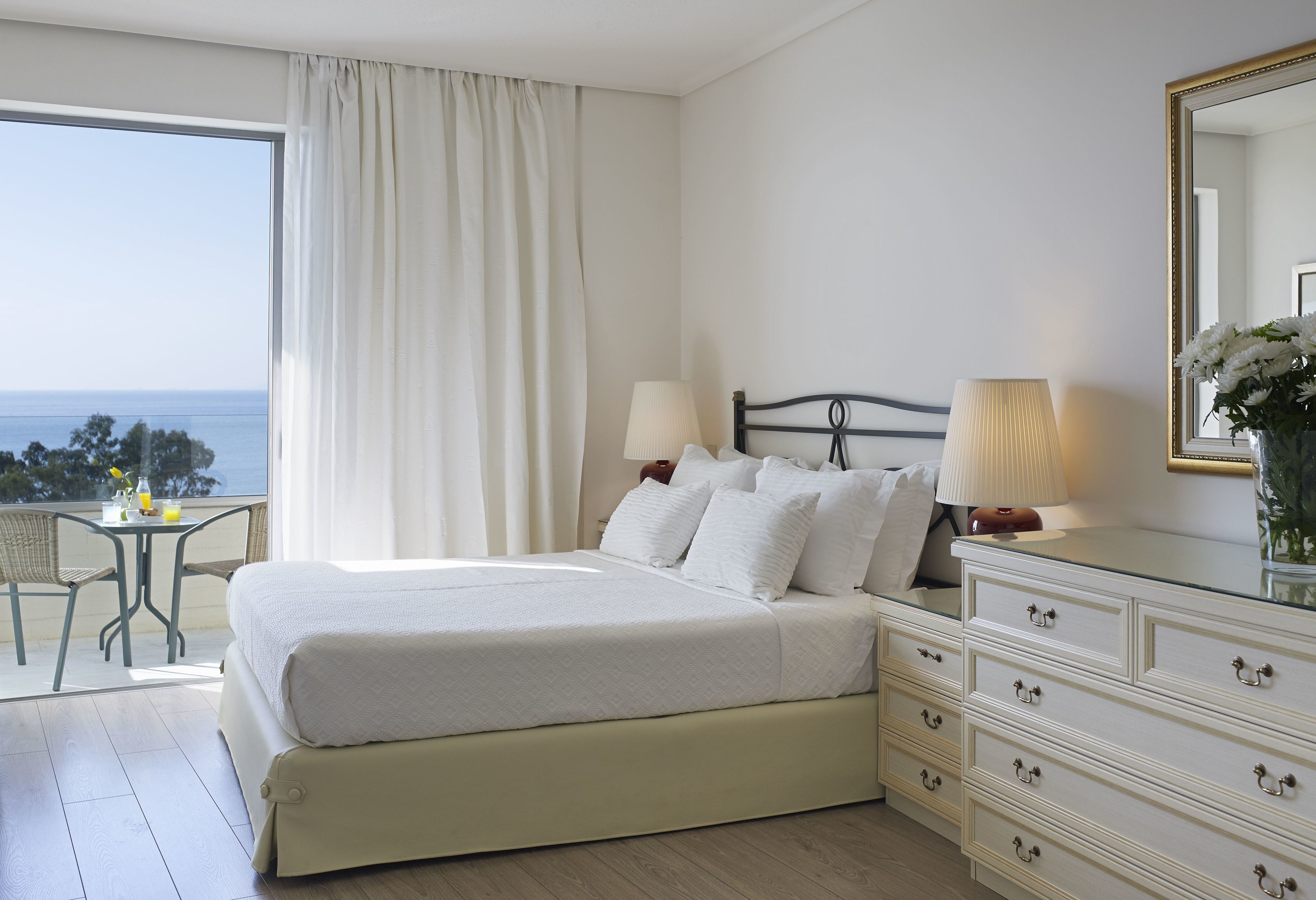 Ein Hotelzimmer mit einem großen Bett, zwei Nachttischen mit Lampen, einer Kommode, einem kleinen Tisch mit einem Stuhl und Meerblick durch ein großes Fenster.
