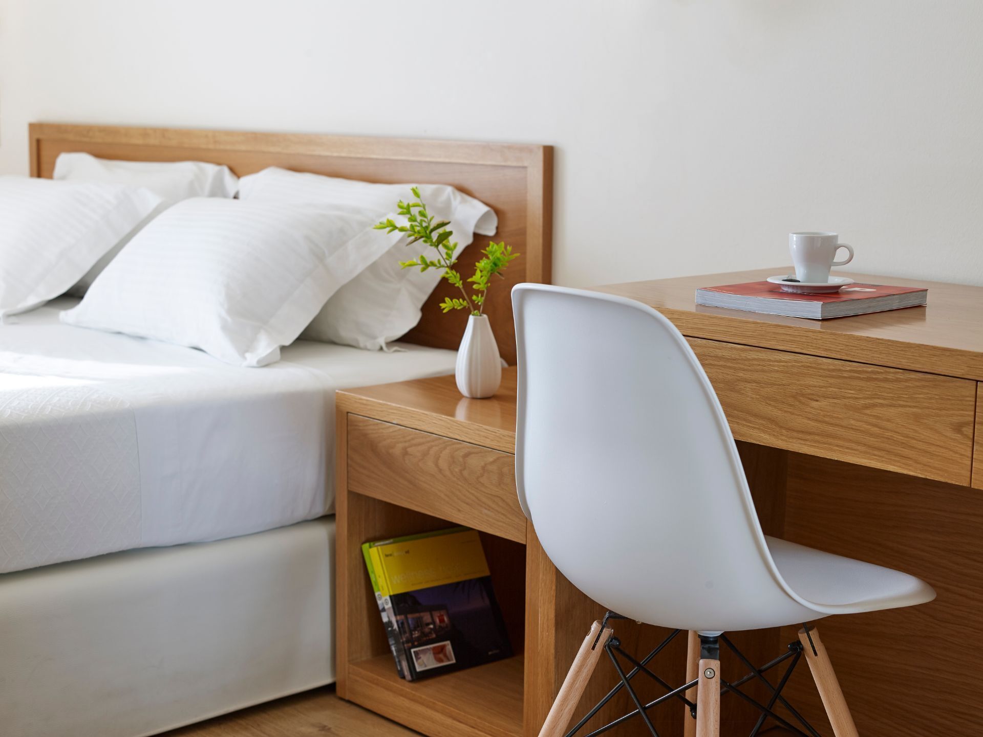 A neatly arranged bedroom with a bed, a bedside table with a book and a cup, and a chair with a plant.