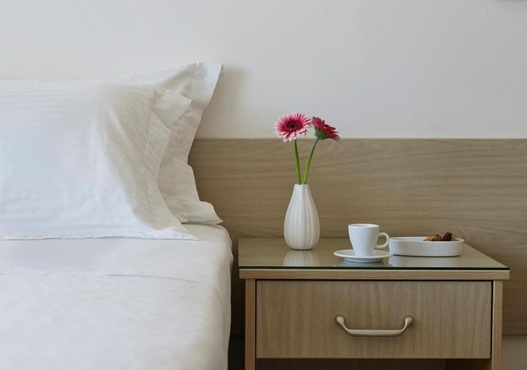 Ein ordentlich bezogenes Bett mit weißen Leinen, ein Nachttisch mit einer weißen Vase mit Blumen, eine weiße Tasse und ein kleiner Teller mit Keksen.