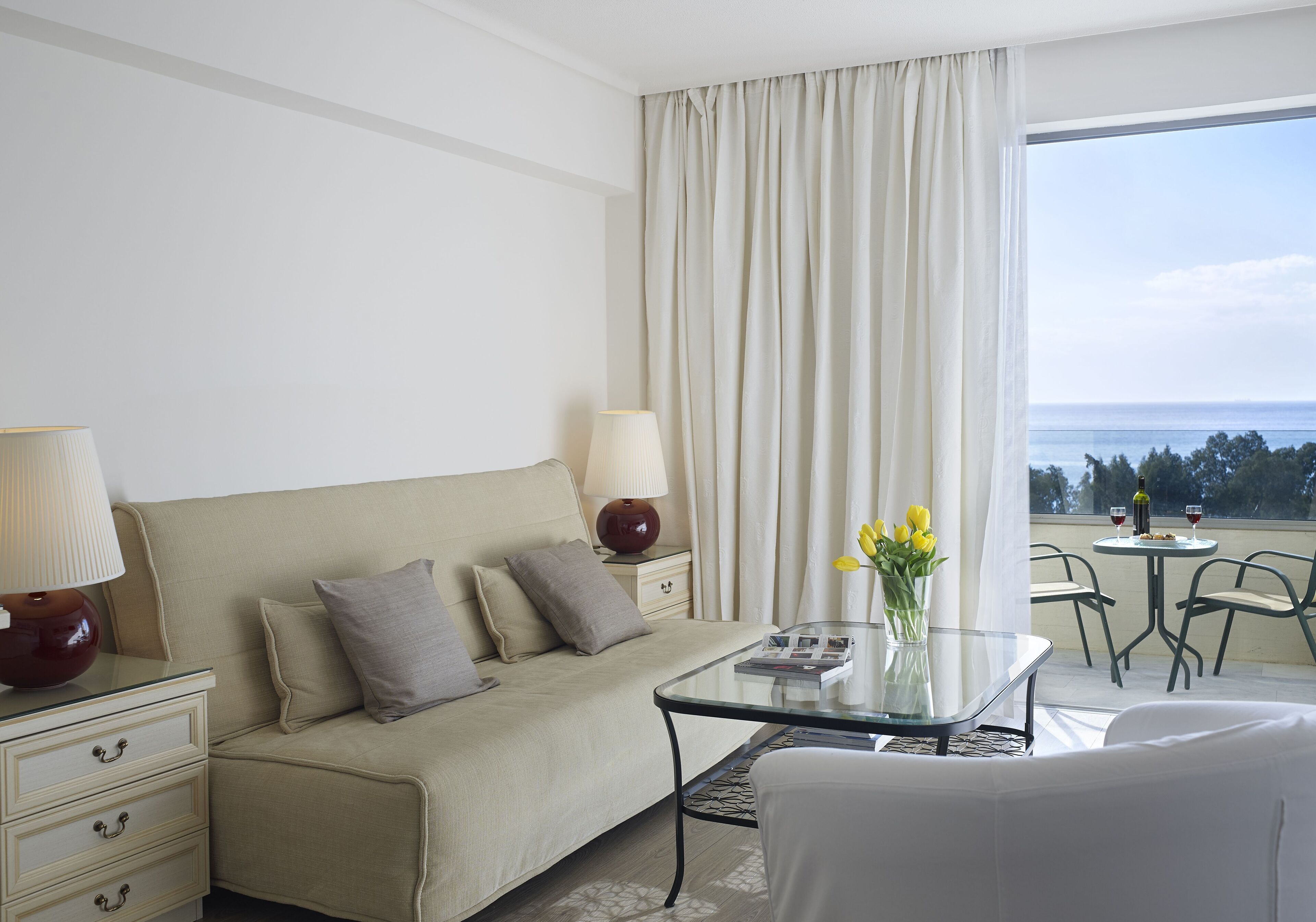 A hotel room with a bed, a glass coffee table, a sofa, a small table with a vase of flowers, and a balcony with a view of the sea.