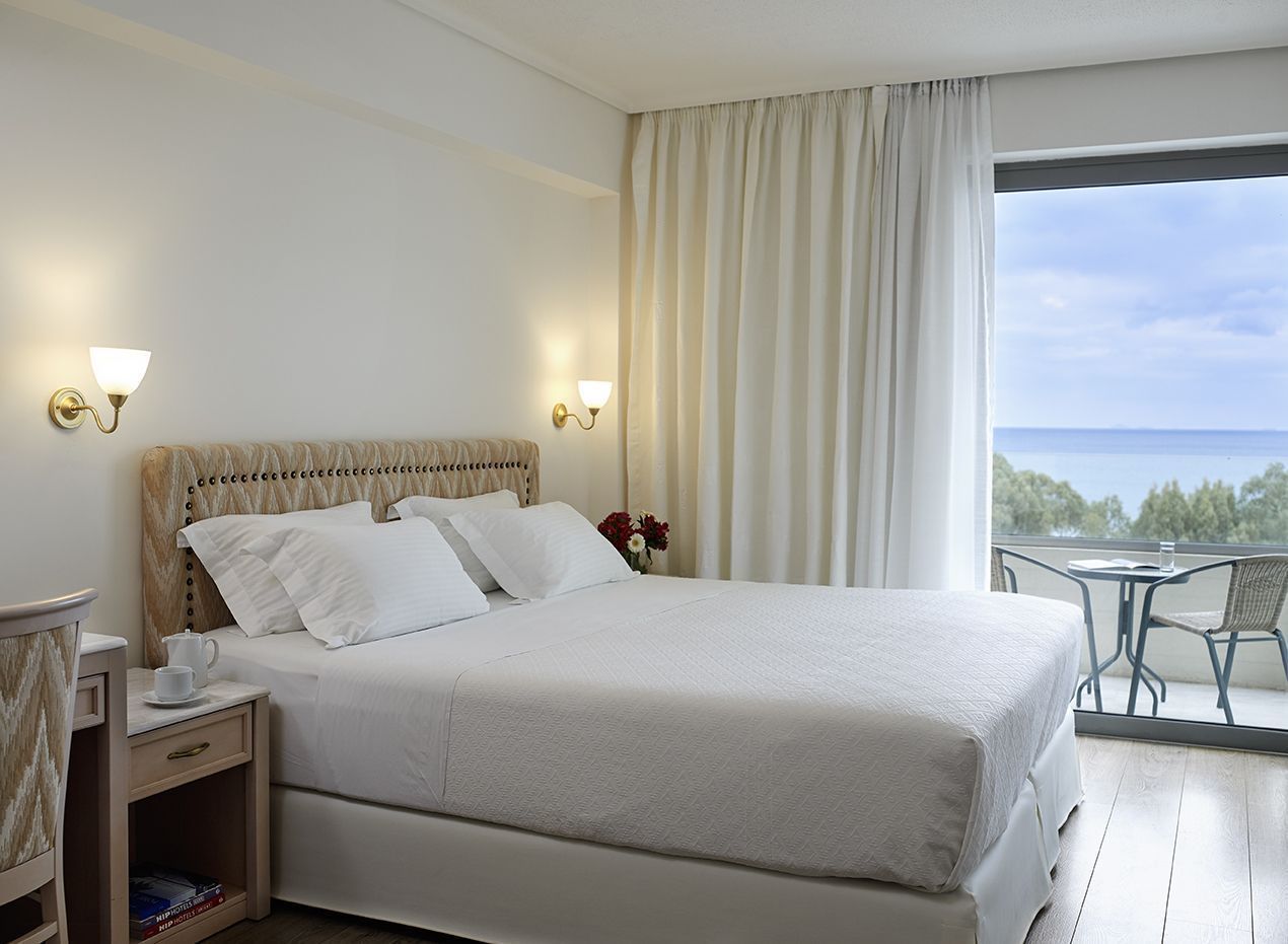 A hotel room with a large bed, bedside tables, a small table with a chair, and a view of the sea through a large window.