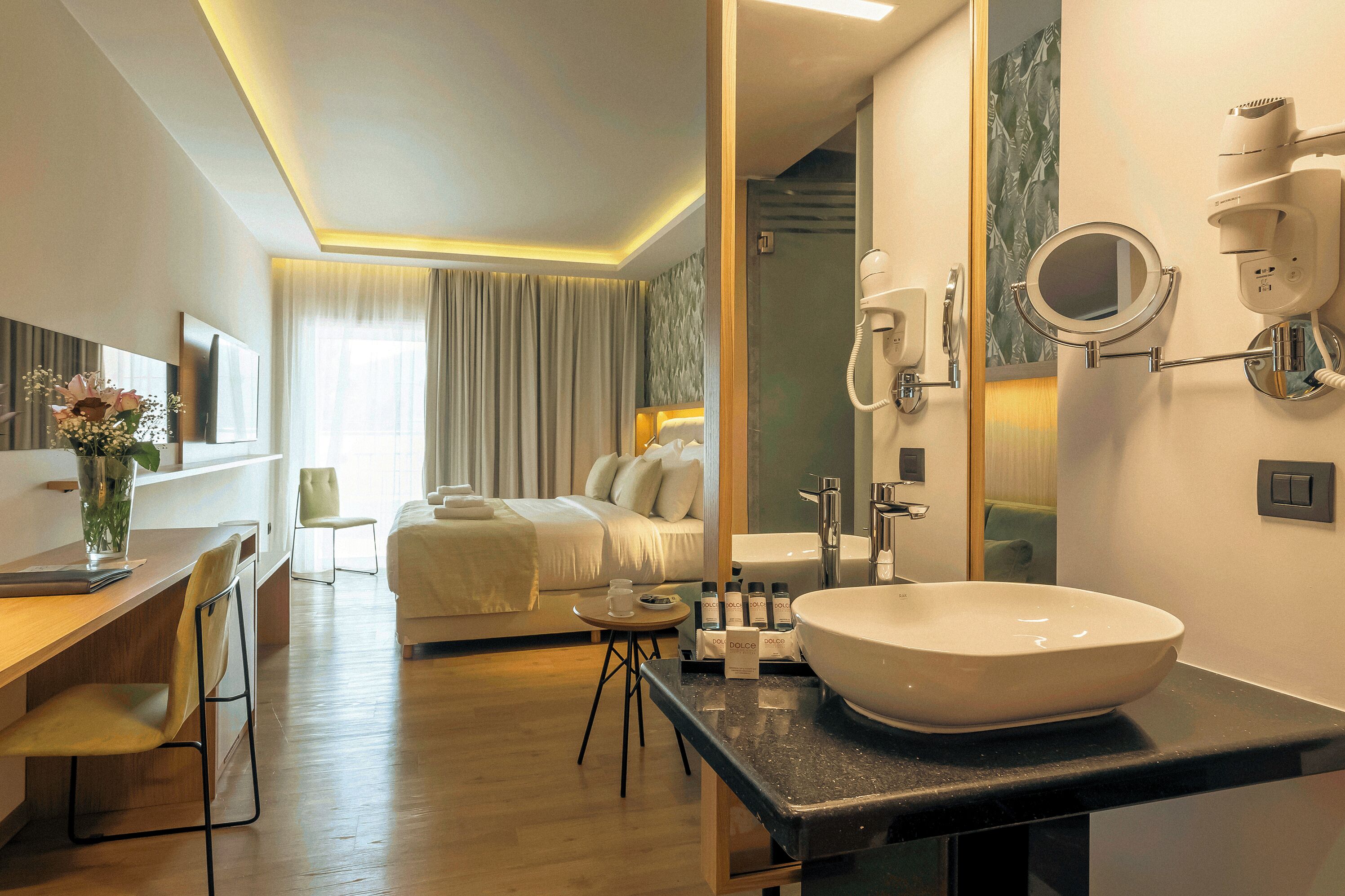 A hotel room with a large bed, a desk with a chair, a round mirror, a sink, and a hairdryer.