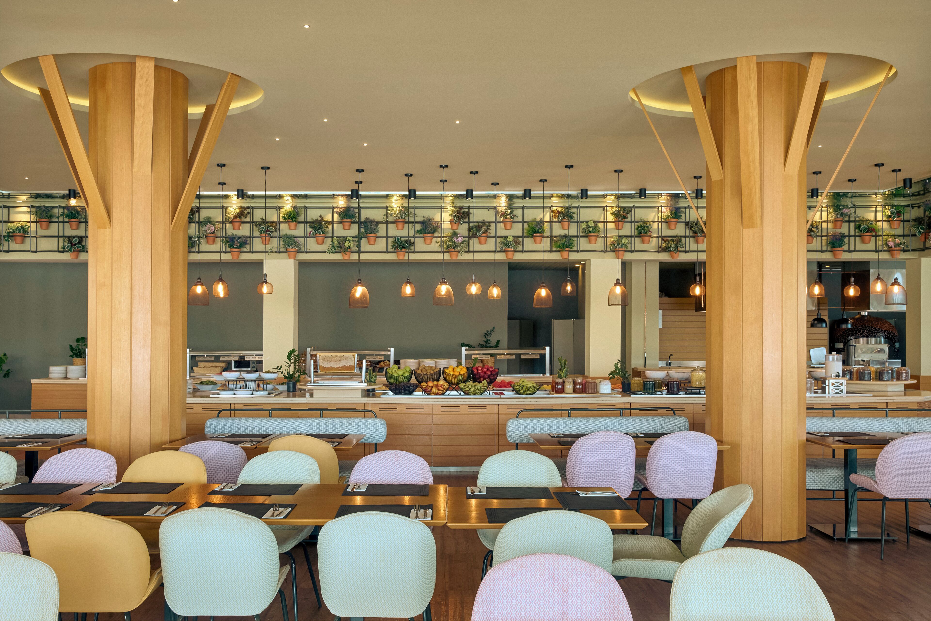 Een modern restaurant met hoge houten pilaren, pastelkleurige stoelen en een centrale balie met een presentatie van fruit.