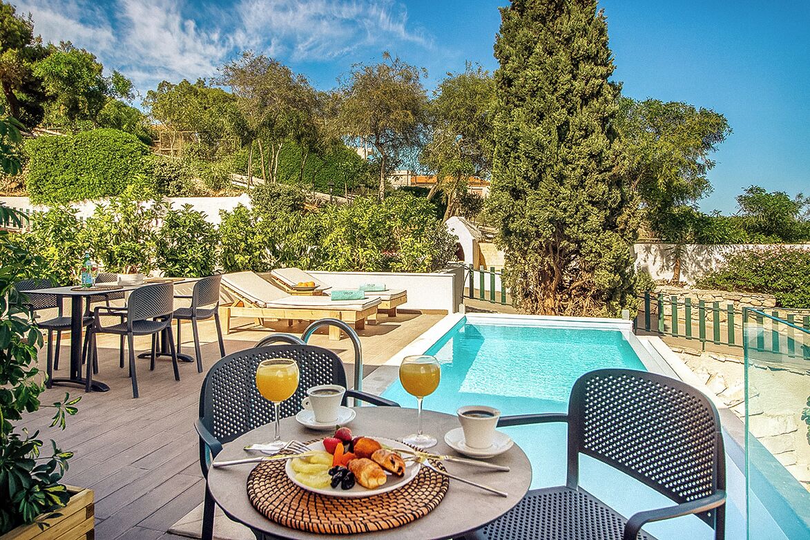 A poolside dining area with a table set for breakfast, including fruits and pastries, and a view of the pool and surrounding greenery.