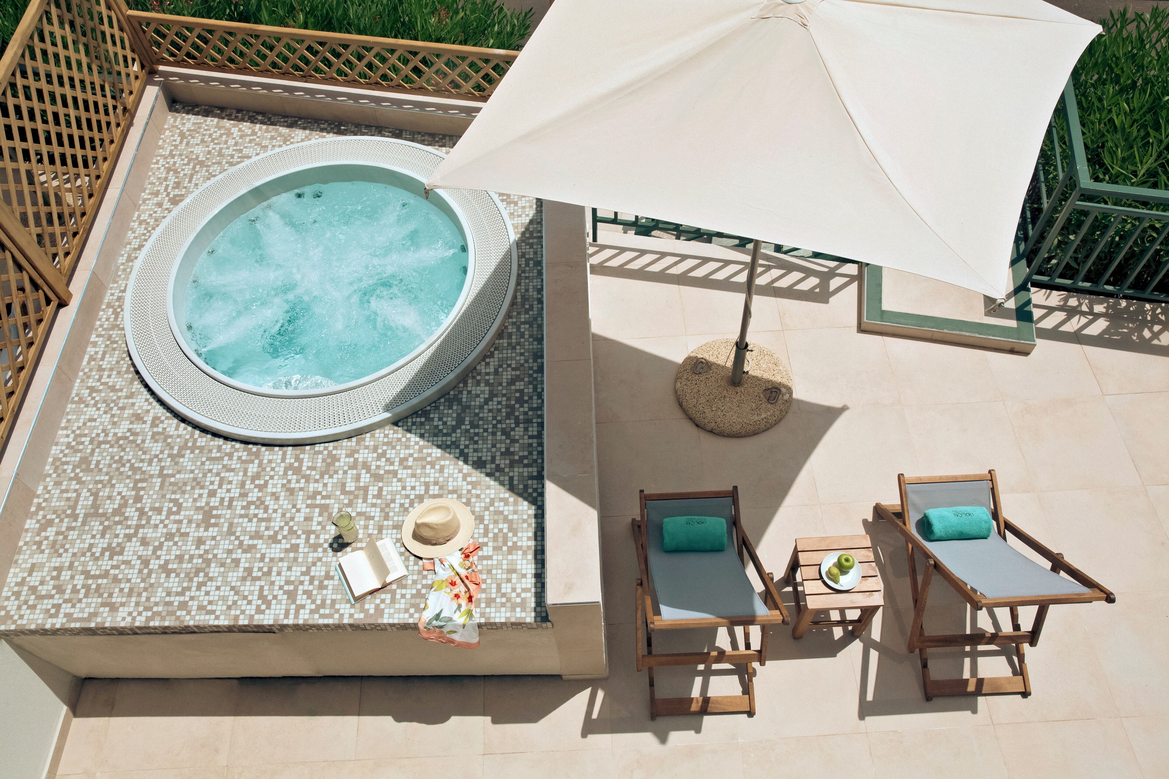 Ein Poolbereich auf dem Dach mit Whirlpool, zwei Liegestühlen, einem Tisch mit Getränk und einem großen Sonnenschirm, der Schatten spendet.