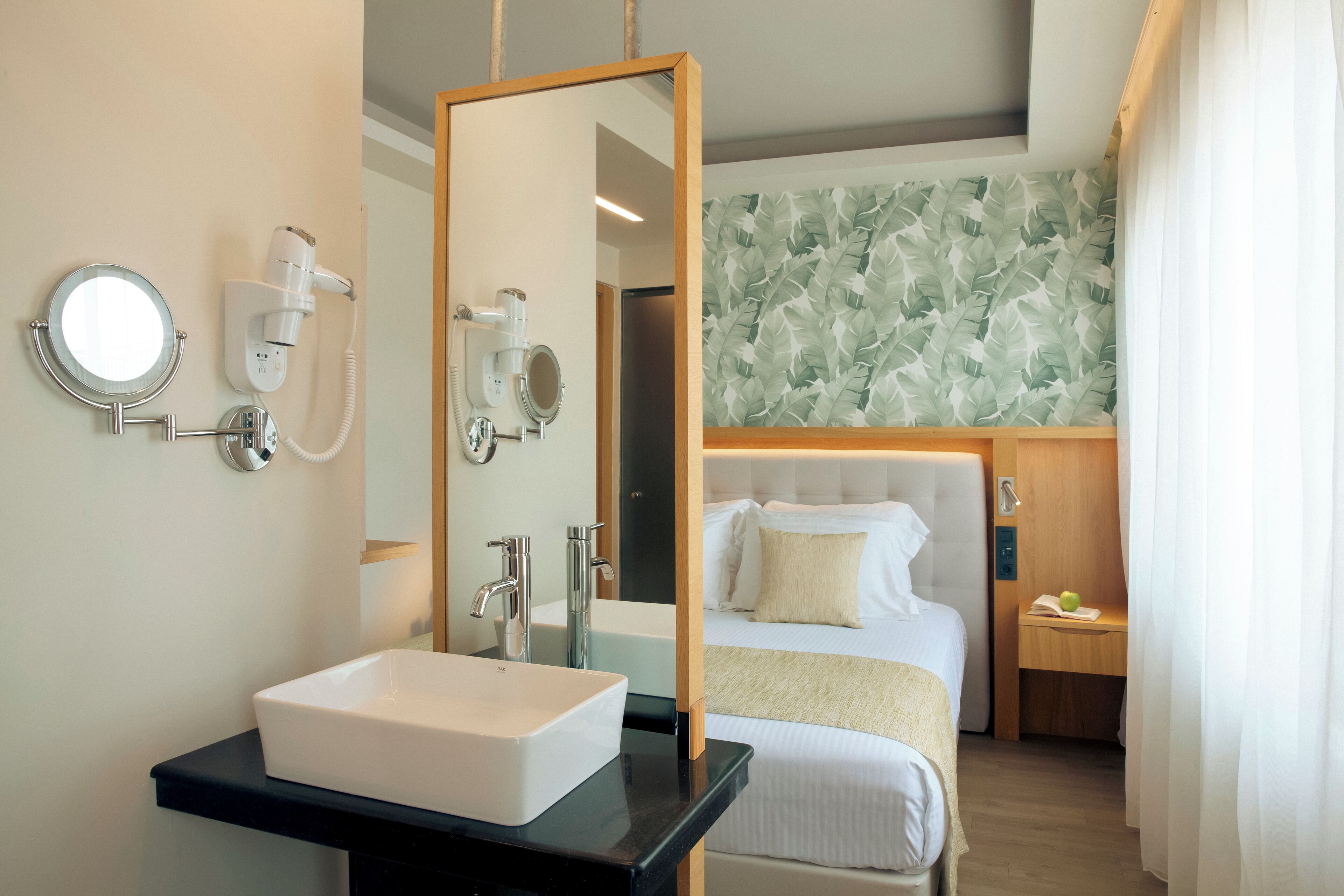 A modern hotel room with a large mirror, a white sink, a bed with a white headboard, and a wooden nightstand with a green apple and a book.
