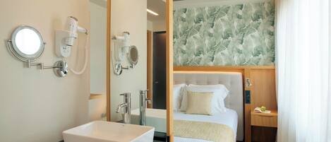 A modern hotel room with a large mirror, a white sink, a bed with a white headboard, and a wooden nightstand with a green apple and a book.