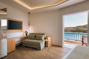 A modern living room with a sofa, a flat-screen TV, a small table, and a balcony with a view of the sea.