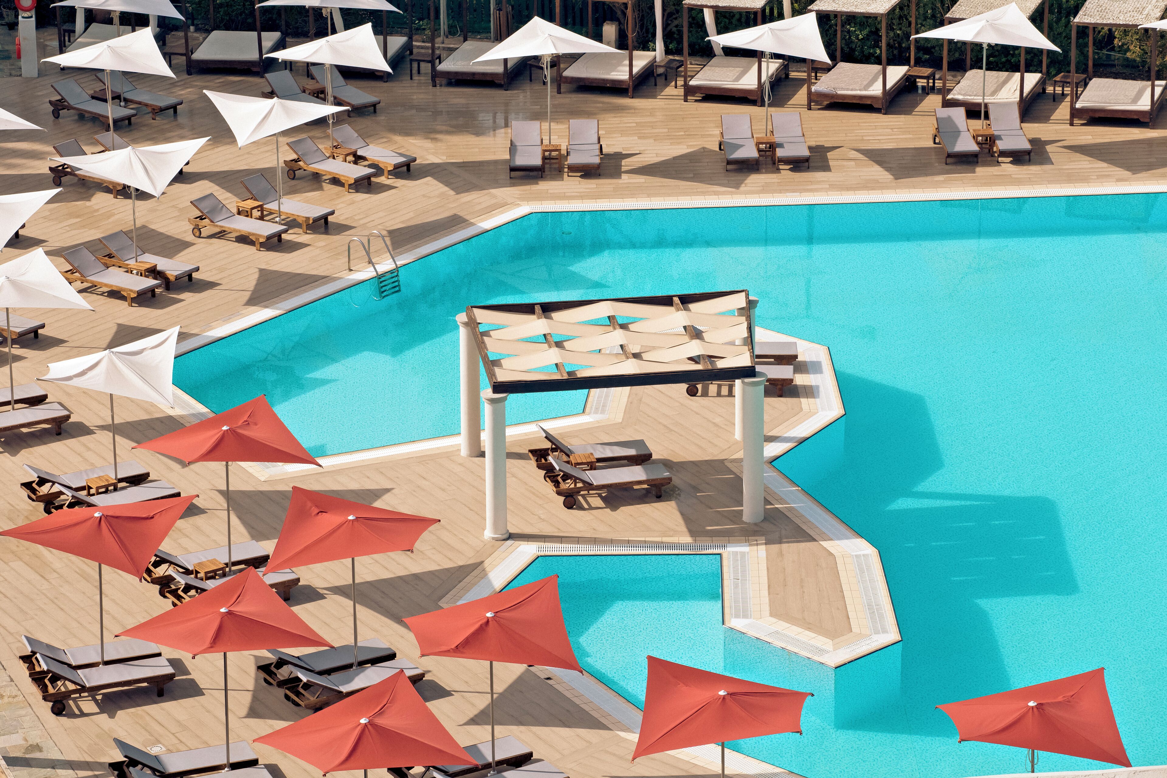 Un área de piscina con múltiples tumbonas y sombrillas, una terraza de madera y una piscina azul cristalina.