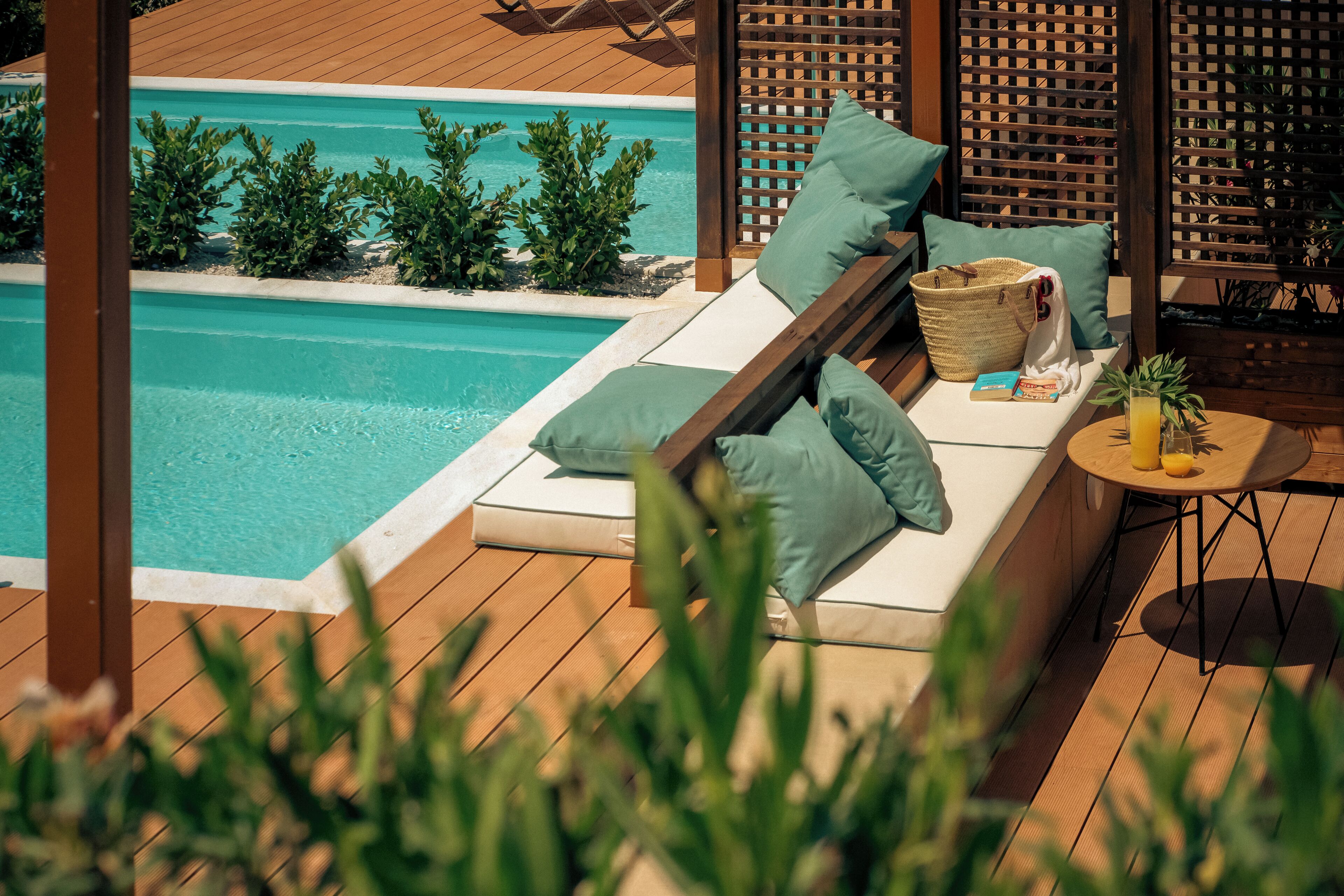 A poolside lounge with a wooden deck, a cushioned seating area with pillows, a small table with a drink, and a basket.