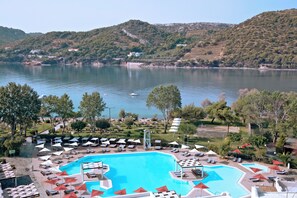 A pool area with lounge chairs and umbrellas, surrounded by greenery and a view of a lake. - Zeus Dolce by Wyndham Athens (Markopoulo Mesogaias)