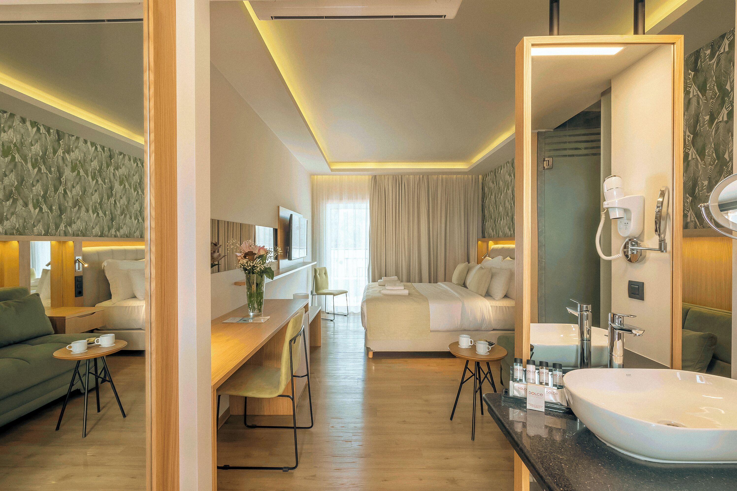 Habitación de hotel con cama, escritorio y silla, sofá, baño con lavamanos y espejo, y ducha.