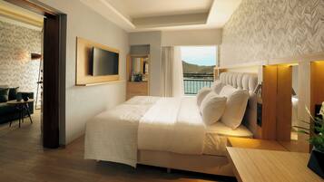 A modern bedroom with a large bed, a TV mounted on the wall, a balcony with a view of the sea, and a wooden desk.