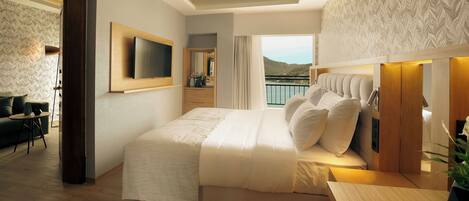Un dormitorio moderno con una cama grande, un televisor montado en la pared, un balcón con vistas al mar y un escritorio de madera.
