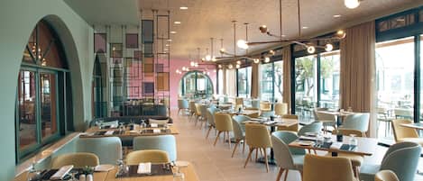 A modern restaurant with multiple dining tables and chairs, large windows, and a patterned ceiling.