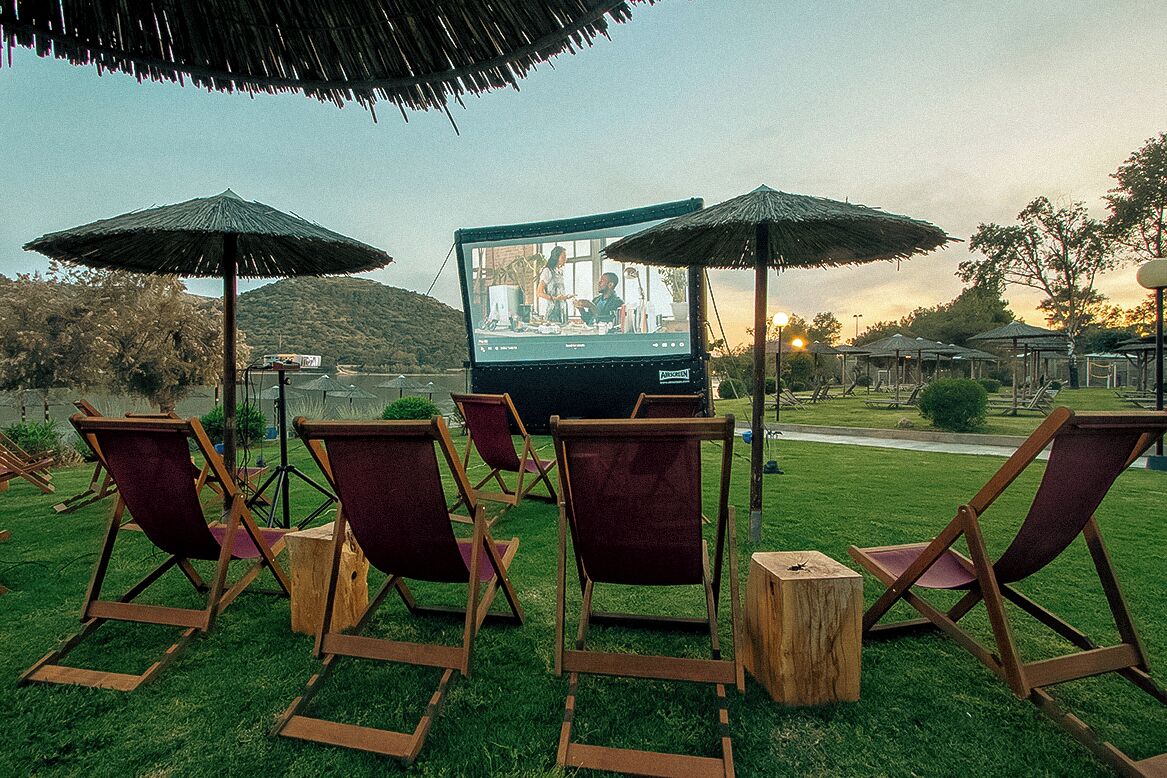 Cine al aire libre con una pantalla grande, sillas de madera y sombrillas de paja.