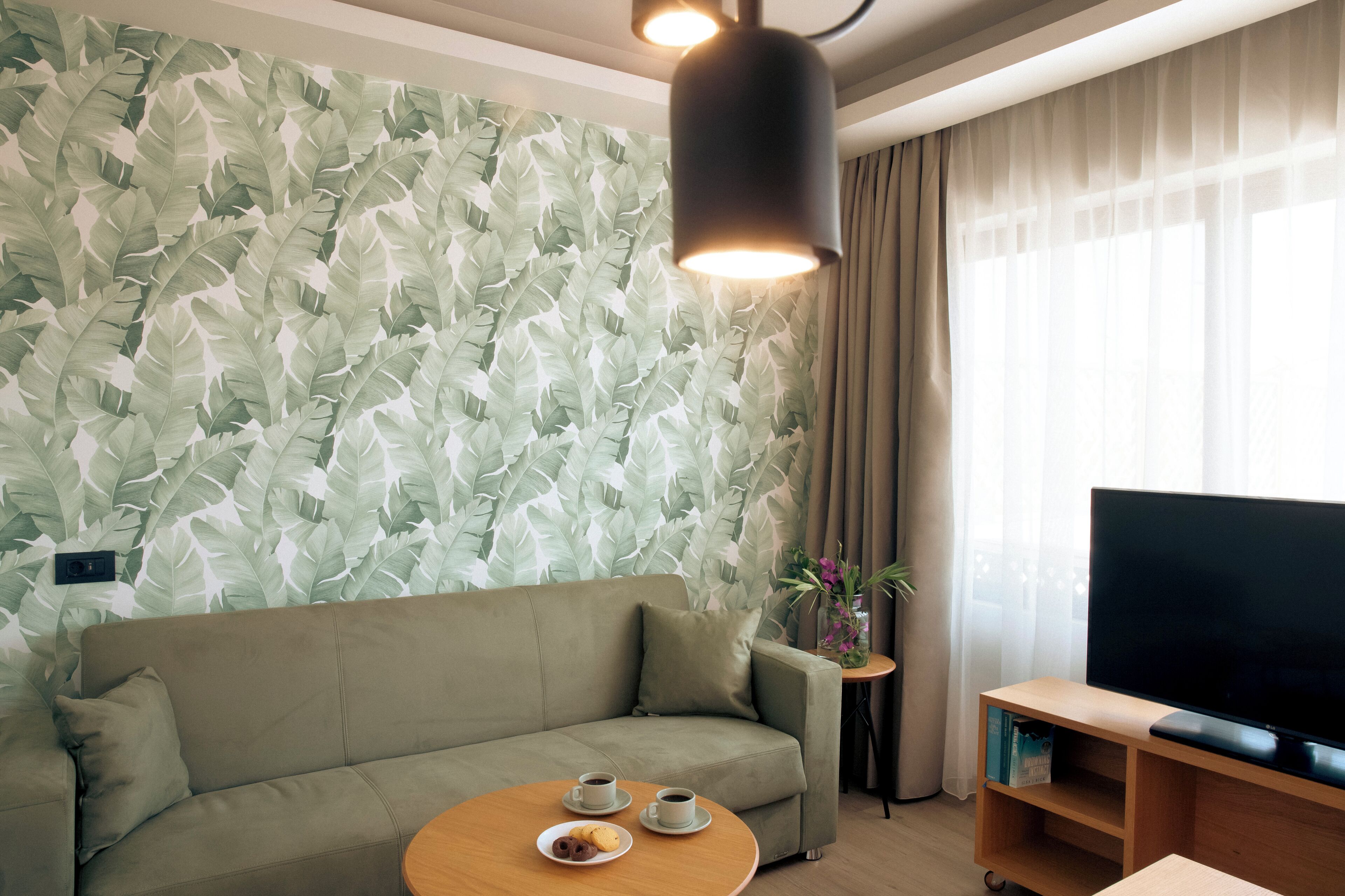 A modern living room with a sofa, a coffee table with cups and pastries, a flat-screen TV, and a wall with a leafy pattern.