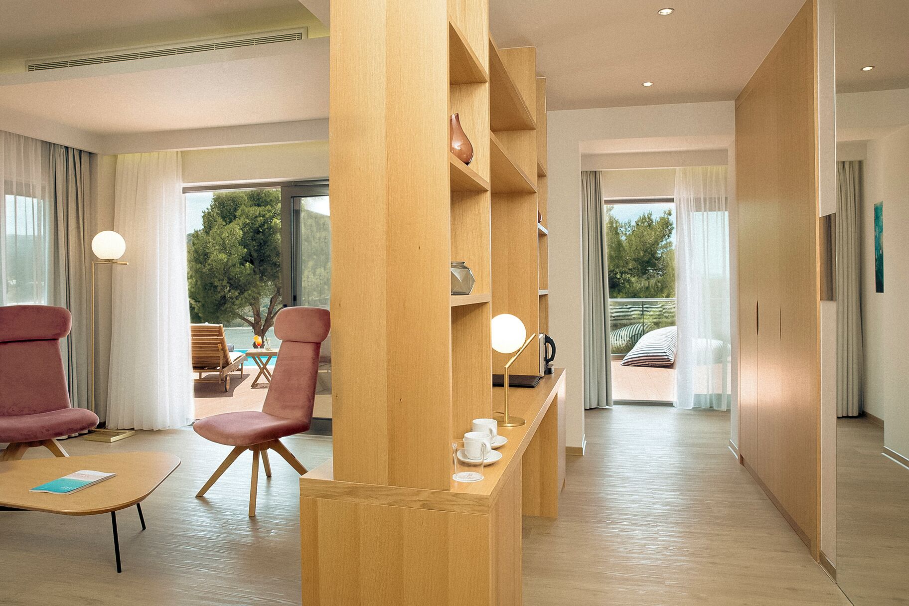 Een moderne hotelkamer met een houten receptiebalie, een zithoek met een roze stoel, en uitzicht op het landschap door grote ramen.