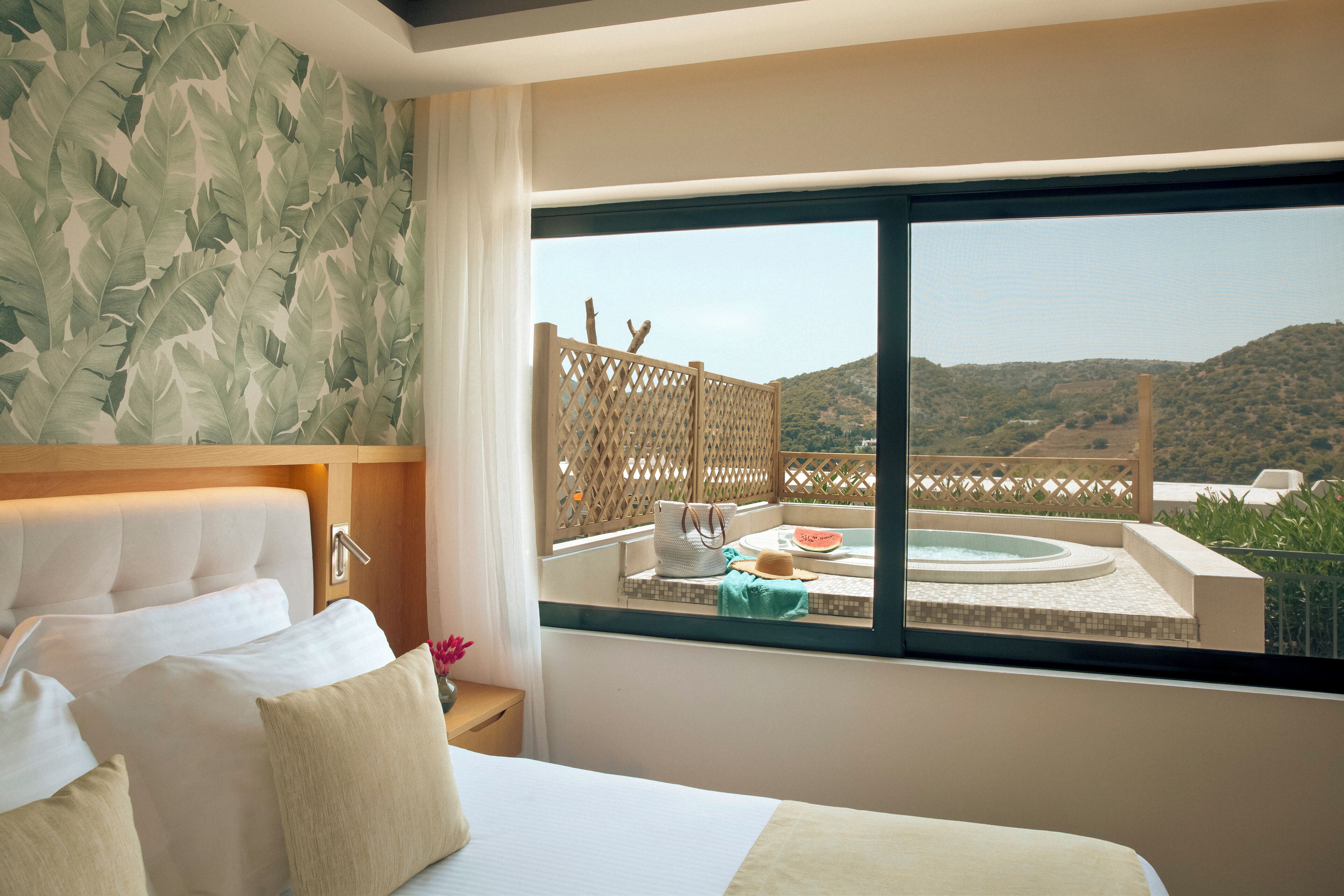 A hotel room with a large bed, a view of a hot tub and mountains, and a tropical leaf wallpaper.