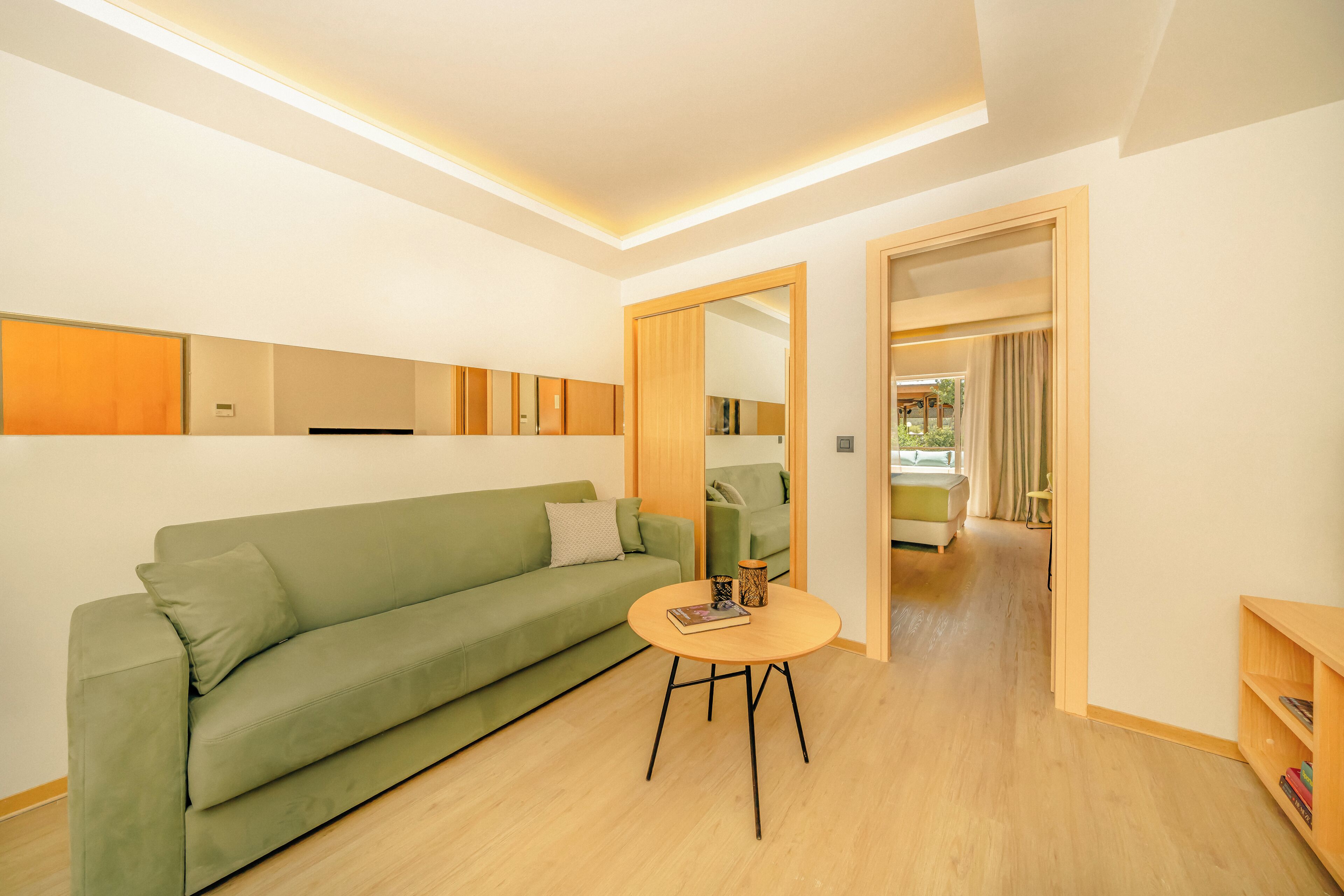 A modern living room with a green sofa, a round wooden coffee table, and a mirror reflecting another room.