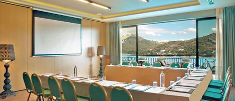 A conference room with a long table, chairs, a projector screen, and a view of a mountainous landscape through large windows.