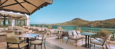 An outdoor terrace with tables and chairs, overlooking a scenic landscape with hills and a body of water.