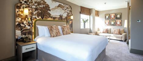 Una cama grande con cabecera, una mesita de noche con lámpara, una pequeña mesa y un sofá en una habitación luminosa con vistas al exterior.