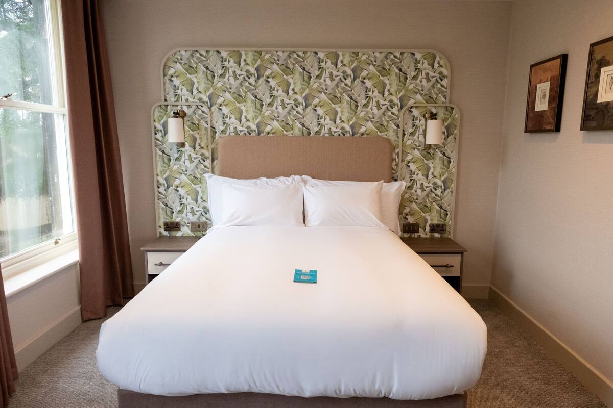 A hotel room with a large bed, two bedside tables, a window with curtains, and a wall with framed pictures.