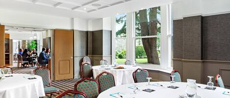 Une salle de conférence dotée de tables rondes, de chaises à motifs turquoise et de grandes fenêtres offrant une vue sur les arbres.