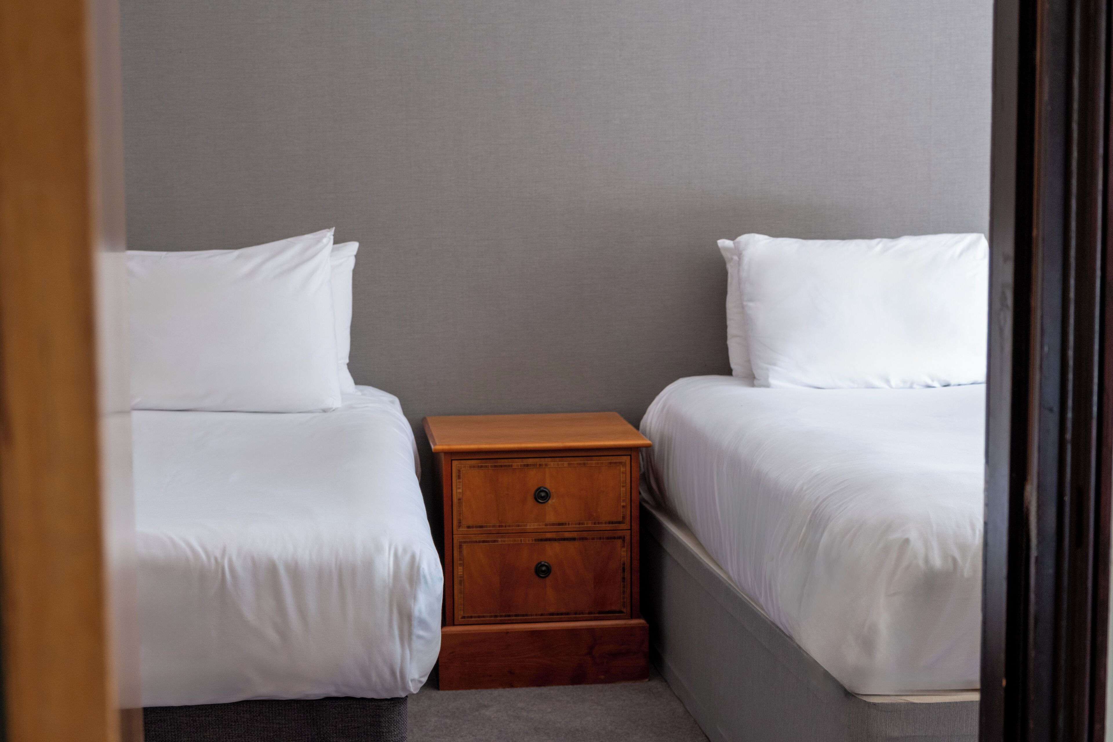 Un dormitorio pequeño y ordenado con dos camas individuales, una mesita de noche de madera y una pared gris.