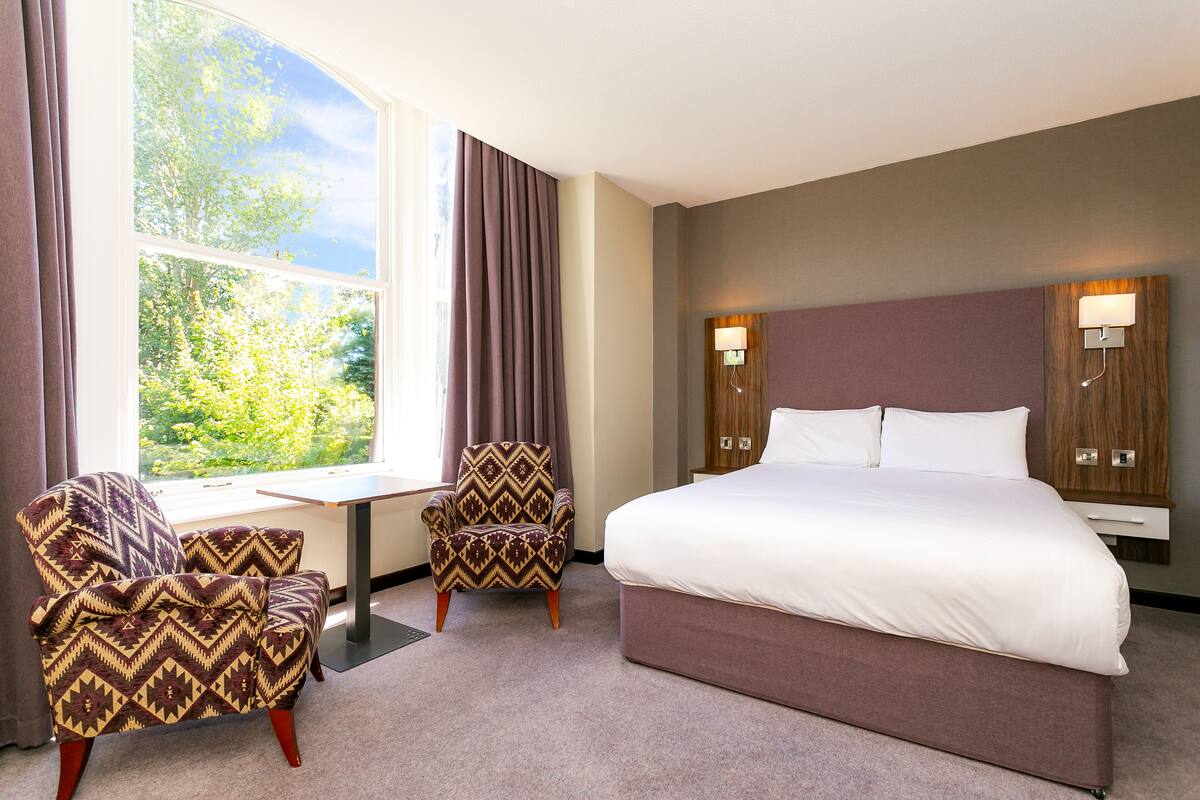 A hotel room with a large bed, two armchairs, a small table, and a window with curtains.