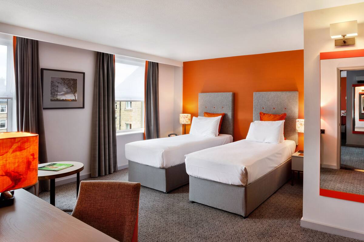 A hotel room with two beds, a desk, a chair, a lamp, a framed picture, and a window with curtains.