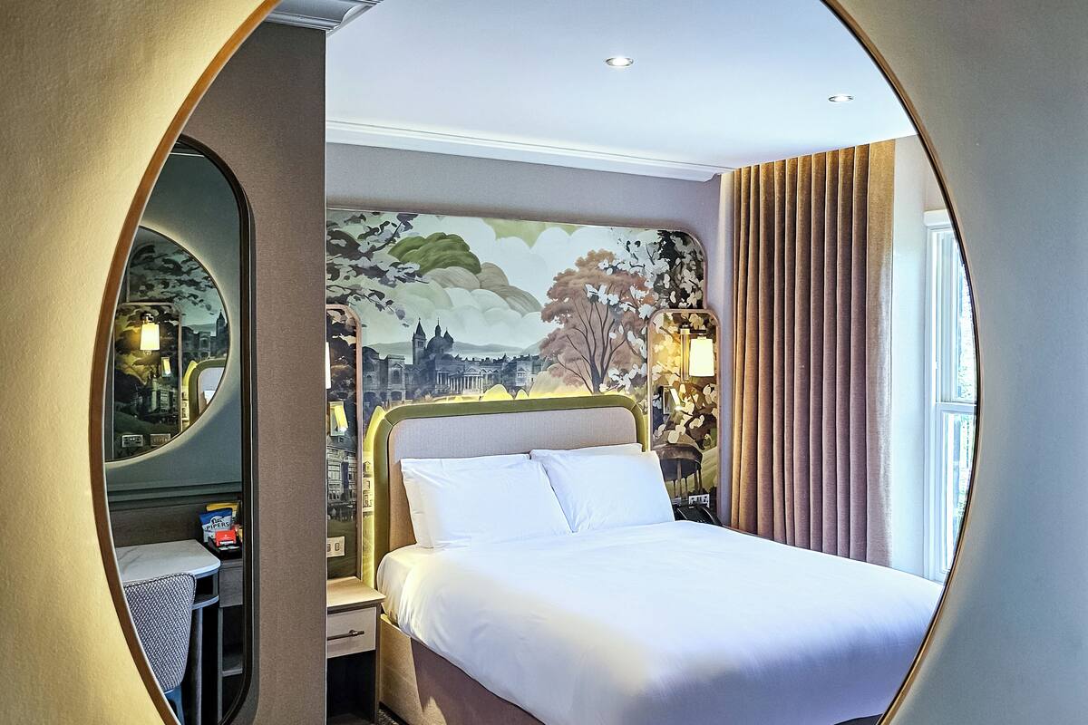 A hotel room with a large round mirror, a bed with white linens, a nightstand, a lamp, and a mural on the wall.