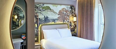 A hotel room with a large round mirror, a bed with white linens, a nightstand, a lamp, and a mural on the wall.