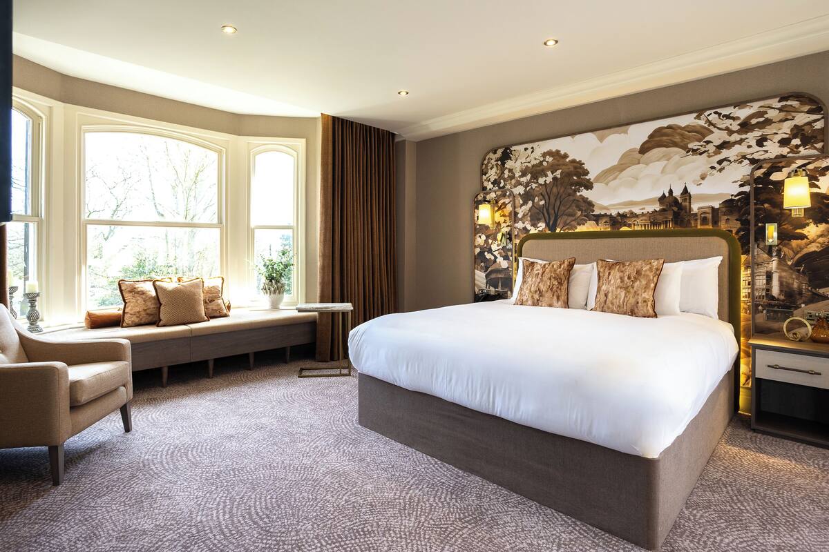 A spacious bedroom with a large bed, a seating area by the window, and a decorative headboard.